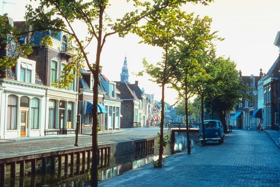 Straße entlang eines Kanals mit Bäumen, historische Gebäude und ein Auto.