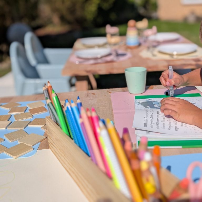 Bunte Buntstifte und ein Kind, das im Freien auf Papier malt. Tisch mit Geschirr im Hintergrund.