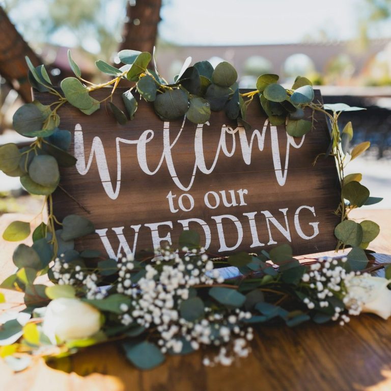 Willkommensschild für eine Hochzeit, umgeben von grünen Blättern und Blüten.