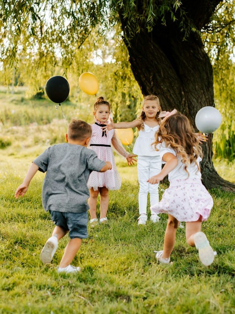 Vier Kinder spielen mit Luftballons unter einem Baum auf einer Wiese.