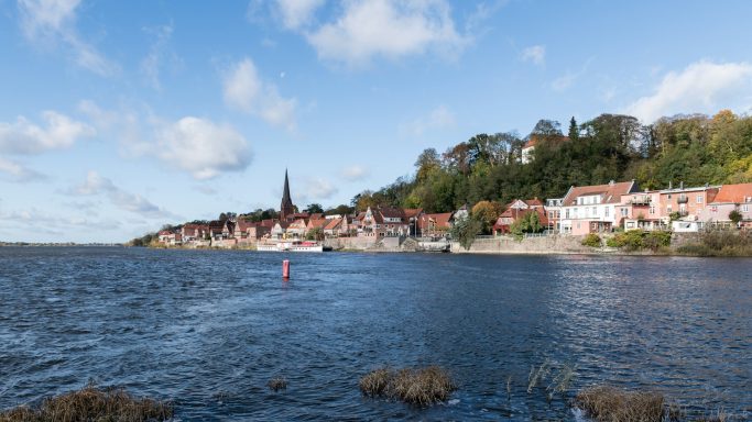 Landschaft mit einem See, einer Stadt am Ufer und bewaldeten Hügeln im Hintergrund.