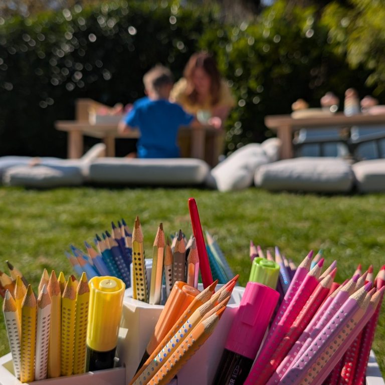 Bunte Stifte in einem Behälter, mit Kindern und einer Frau im Hintergrund beim Spielen.
